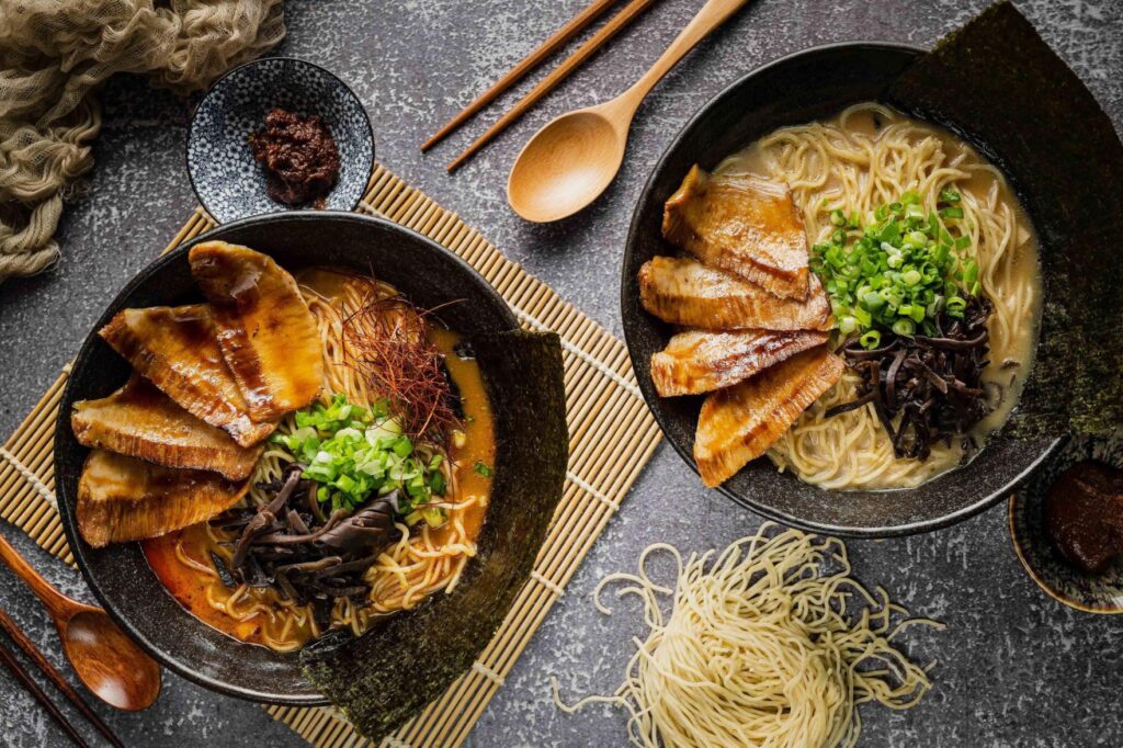 Two bowls of ramen are artfully presented on a textured grey surface, topped with sliced chashu pork, scallions, wood ear mushrooms, and crisp nori sheets. The scene is complemented by wooden utensils, small side dishes of chili paste, and a bundle of dry noodles for a rustic, culinary aesthetic.