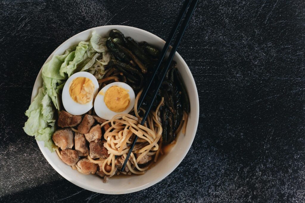 A white bowl filled with ramen noodles, sliced hard-boiled eggs, meat chunks, and greens is centered against a dark, textured background. Black chopsticks rest across the noodles, which are served in a savory dark broth.