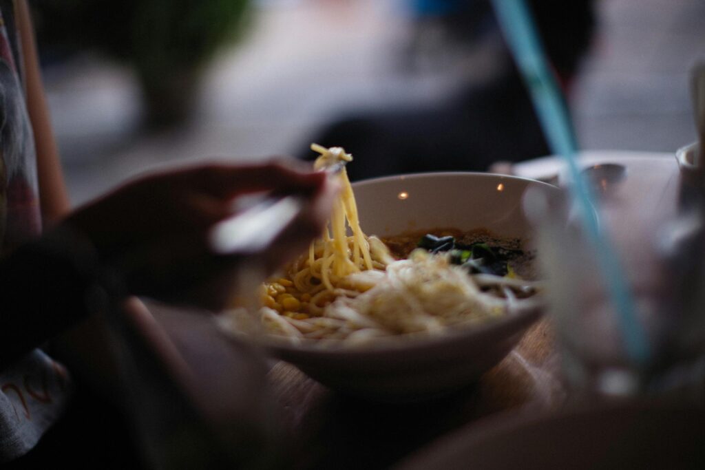 A person uses chopsticks to lift a steaming portion of noodles from a bowl filled with corn and bean sprouts. The scene is captured with a shallow depth of field, focusing on the meal while the surrounding outdoor environment remains softly blurred.