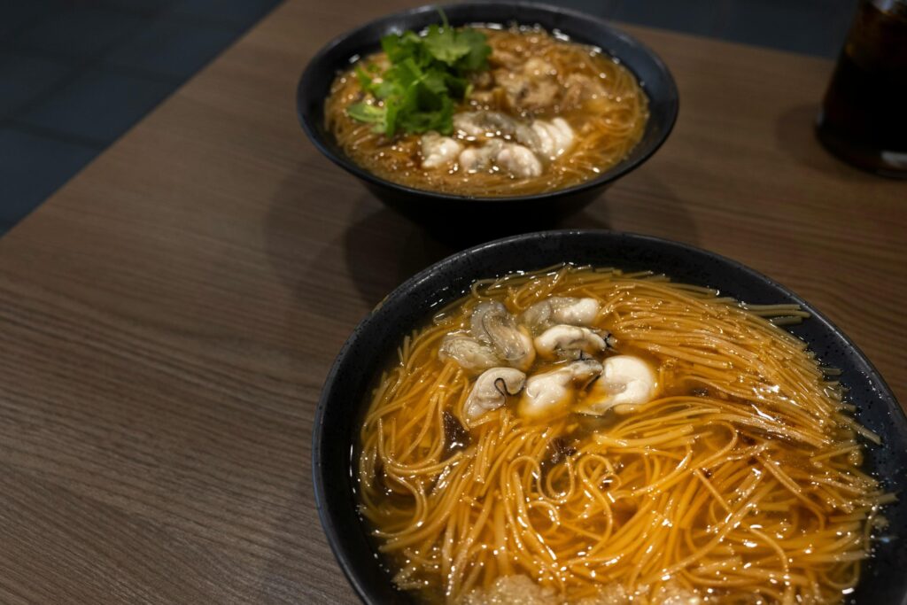 Two bowls of Taiwanese oyster vermicelli sit on a dark wooden table, featuring thin noodles in a savory, thickened broth. The dish is topped with plump, poached oysters and garnished with fresh green cilantro.