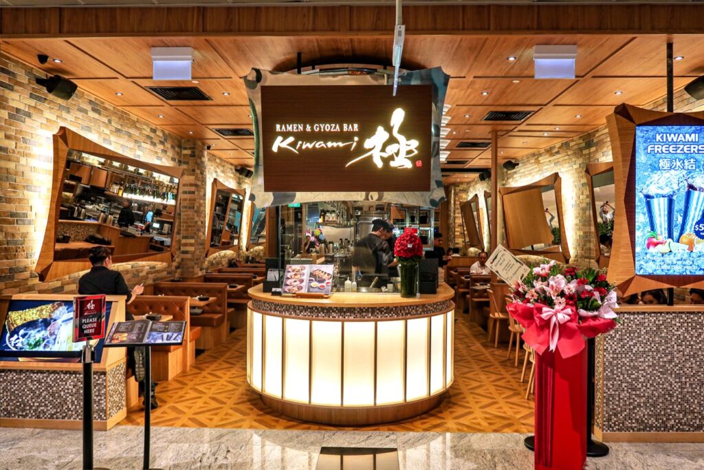 The entrance to Kiwami Ramen & Gyoza Bar features a bright, curved service counter and warm wooden paneling accented by brick walls. The interior is furnished with cozy booth seating and decorated with celebratory floral stands near the "Please Queue Here" sign.