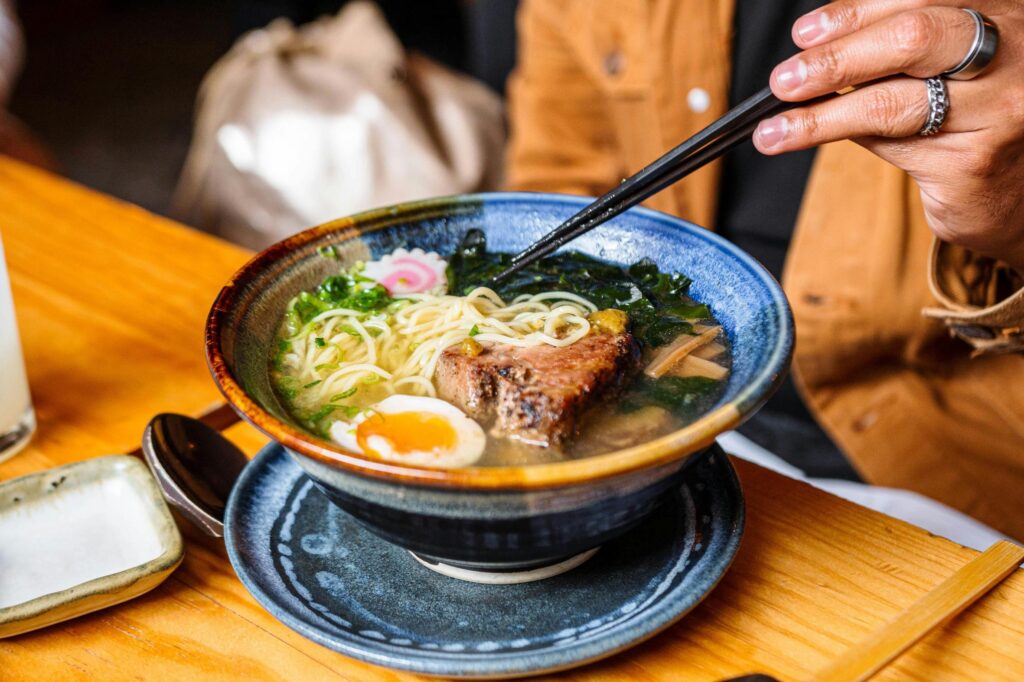 A person wearing rings uses black chopsticks to lift noodles from a blue ceramic bowl of ramen filled with pork, a soft-boiled egg, and seaweed. The steaming dish sits on a wooden table alongside a small side plate and a glass of water.