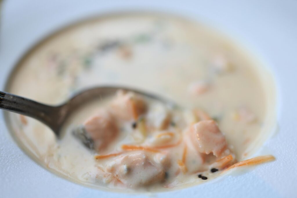 A metal spoon lifts a portion of creamy seafood chowder, revealing chunks of salmon and thin slivers of carrots. The soup is served in a wide white bowl, featuring a rich, opaque broth with hints of herbs and black pepper.
