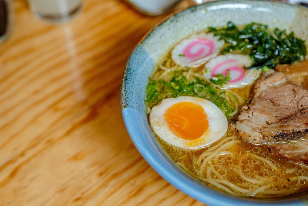 A vibrant bowl of ramen sits on a wooden table, featuring thin noodles and a rich broth topped with a soft-boiled egg and sliced pork. The dish is garnished with colorful pink-swirled narutomaki, fresh green onions, and dark leafy greens.