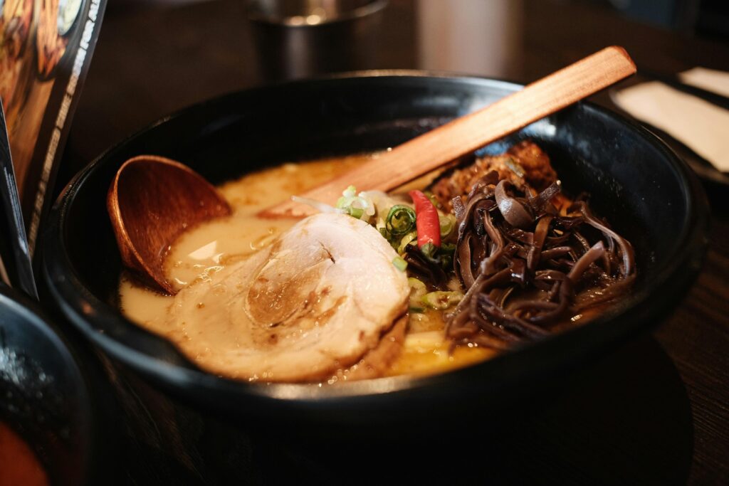 A black bowl of ramen sits on a dark table, featuring a creamy broth topped with a large slice of chashu pork, wood ear mushrooms, and a red chili pepper. A wooden ladle rests inside the bowl, which is surrounded by the soft-focus atmosphere of a restaurant setting.