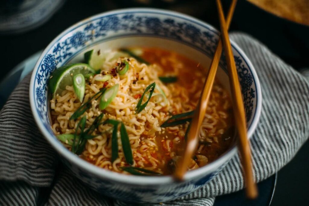 A blue and white patterned bowl is filled with ramen noodles in a spicy reddish broth, topped with sliced scallions, sesame seeds, and a lime wedge. A pair of wooden chopsticks rests across the top of the bowl, which is set against a striped cloth napkin.