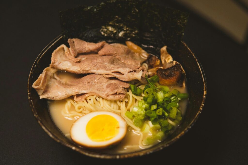 A dark bowl is filled with ramen noodles in a creamy broth, topped with thin slices of meat, a soft-boiled egg, and a generous portion of chopped green onions. A large sheet of dried nori seaweed stands upright against the back of the bowl, which is set against a dark, minimalist background.