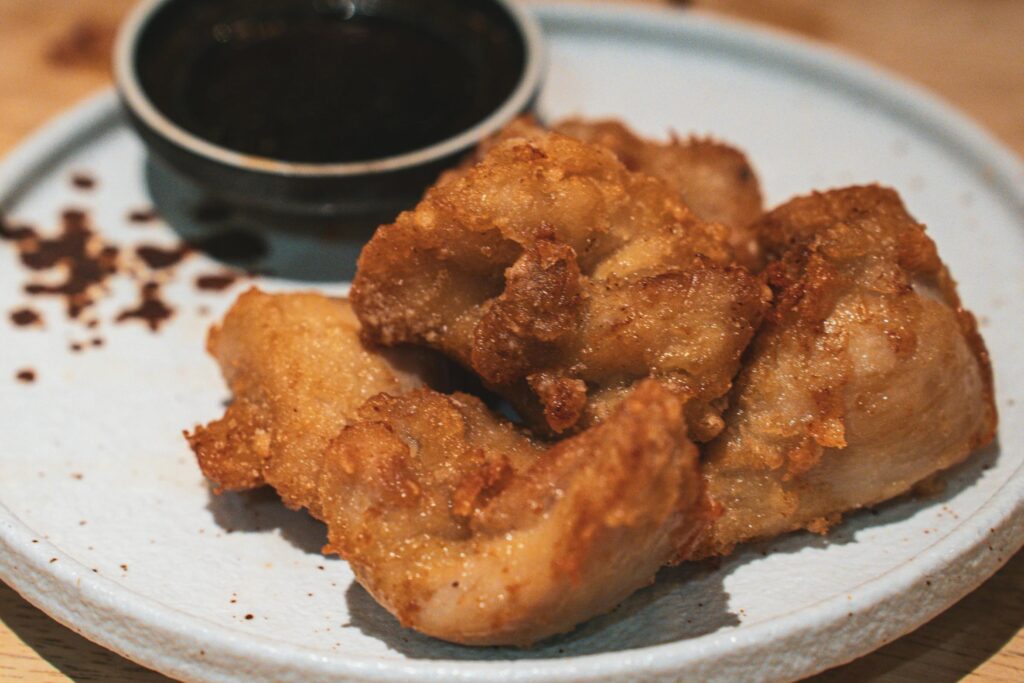 A plate of golden-brown, crispy fried chicken pieces is served on a light-colored ceramic dish. A small black bowl filled with dark dipping sauce sits in the background, accompanied by a light dusting of spice on the side.