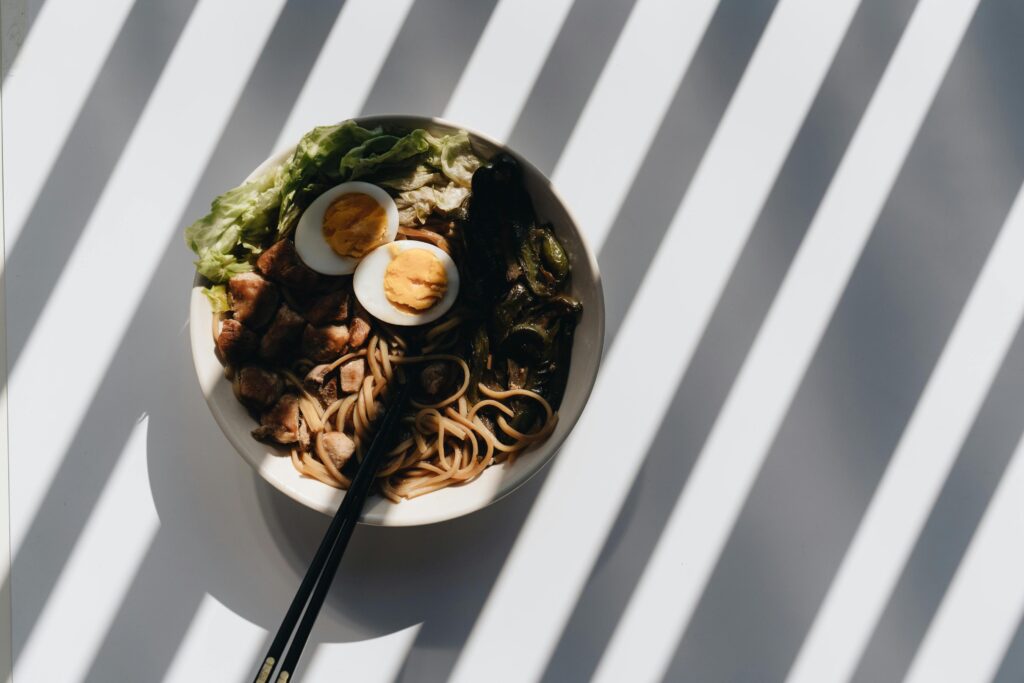 A white bowl of ramen, featuring thick noodles, two hard-boiled egg halves, and chunks of meat, sits on a bright surface. The entire scene is dramatically lit by sharp, diagonal shadows from a nearby window or blind.