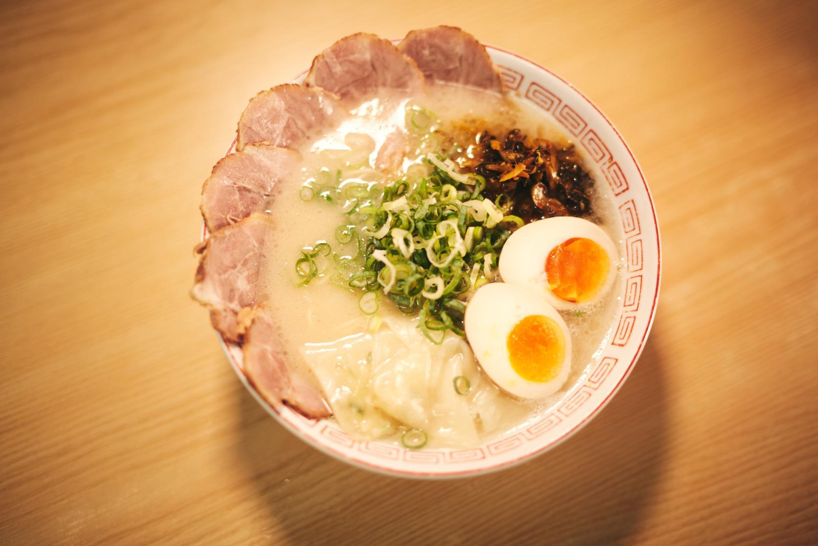 A steaming bowl of tonkotsu ramen is presented on a wooden table, topped with thin slices of chashu pork, soft-boiled eggs, and fresh green onions. The creamy white broth is accented by dark condiments and delicate wontons, all served in a traditional patterned ceramic bowl.