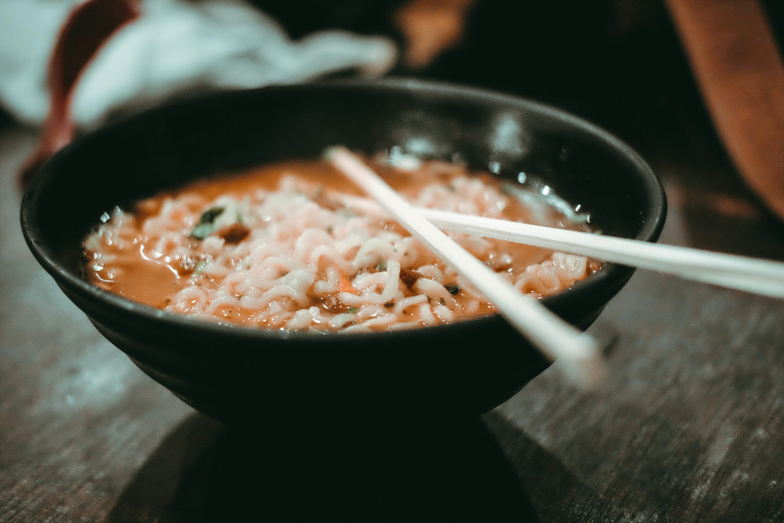 A steaming bowl of ramen is served in a dark ceramic dish with a pair of white chopsticks resting across the rim. The background is softly blurred, keeping the focus on the textured noodles and savory broth.