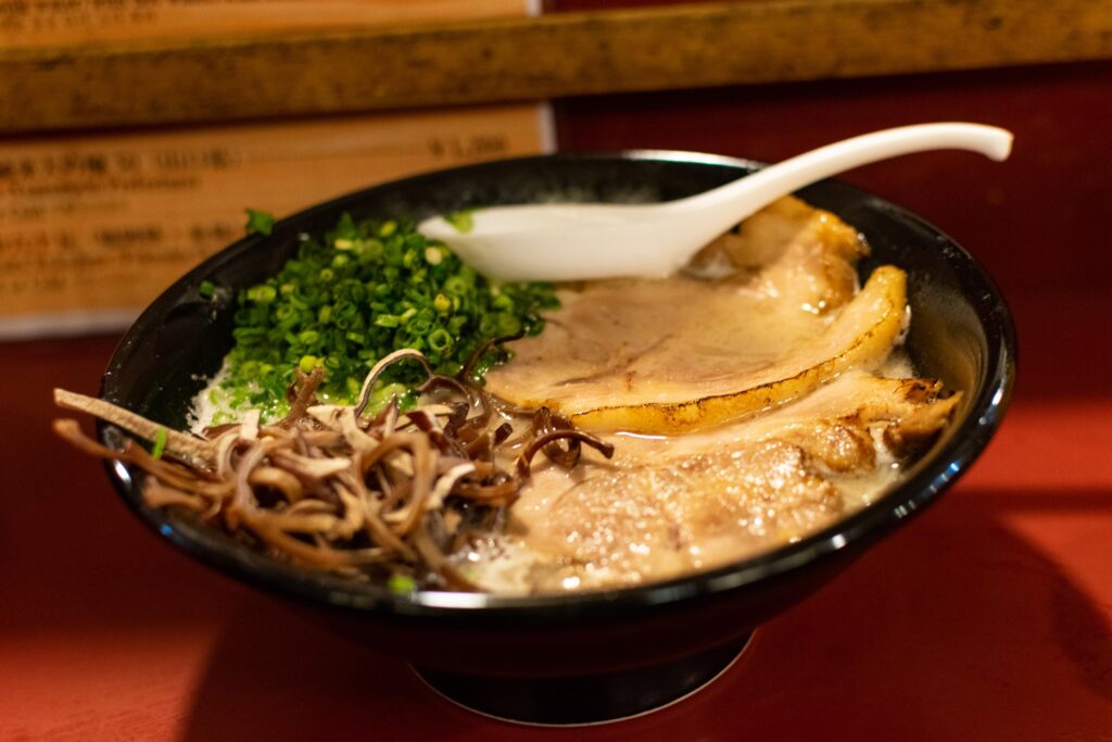 A black bowl filled with rich ramen sits on a red counter, featuring thick slices of chashu pork and a generous pile of chopped green onions. A white ceramic spoon rests in the broth alongside wood ear mushrooms, with a blurred menu visible in the background.