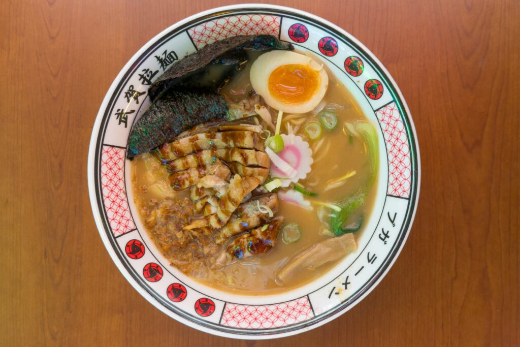 A top-down view shows a steaming bowl of ramen filled with noodles, sliced pork belly, a soft-boiled egg, and nori. The dish is served in a decorative bowl featuring Naruto-inspired Sharingan symbols, resting on a smooth wooden table.