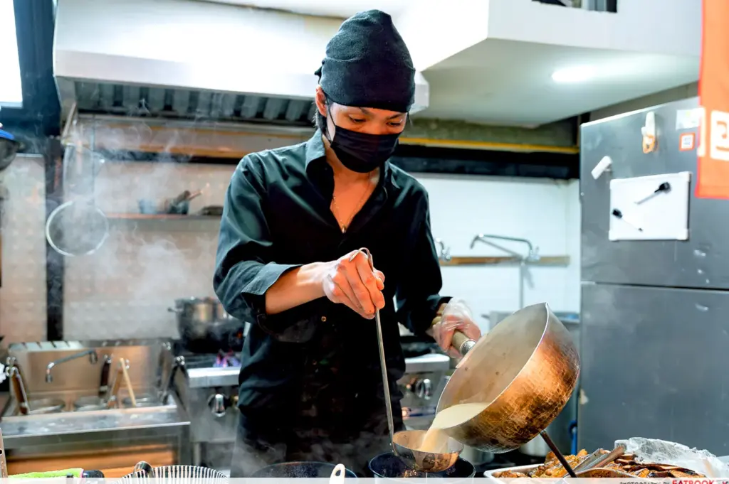 Ramen chef ladling hot broth in a busy kitchen.