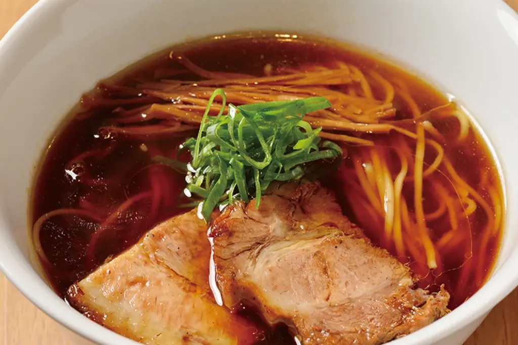 A bowl of Shoyu ramen with a dark savory broth, topped with tender chashu pork slices, bamboo shoots, and fresh green onions.