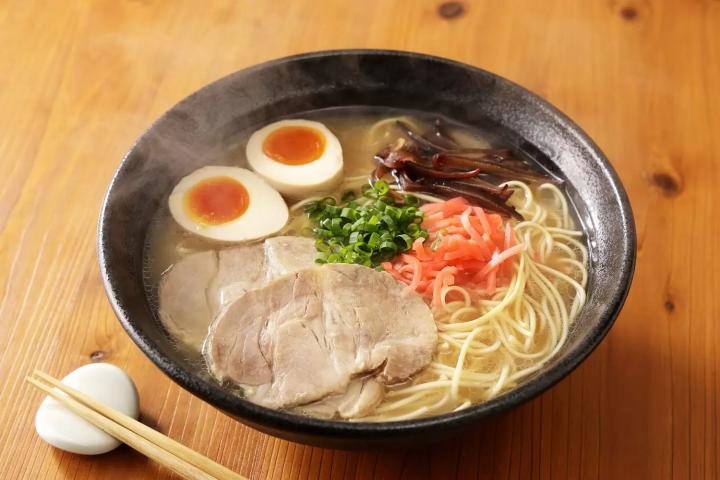 A dark bowl of steaming ramen sits on a wooden table, featuring thin noodles in a clear broth topped with two soft-boiled egg halves and sliced pork chashu. The dish is garnished with bright pink pickled ginger, chopped green onions, and dark wood ear mushrooms, with a pair of chopsticks resting nearby.