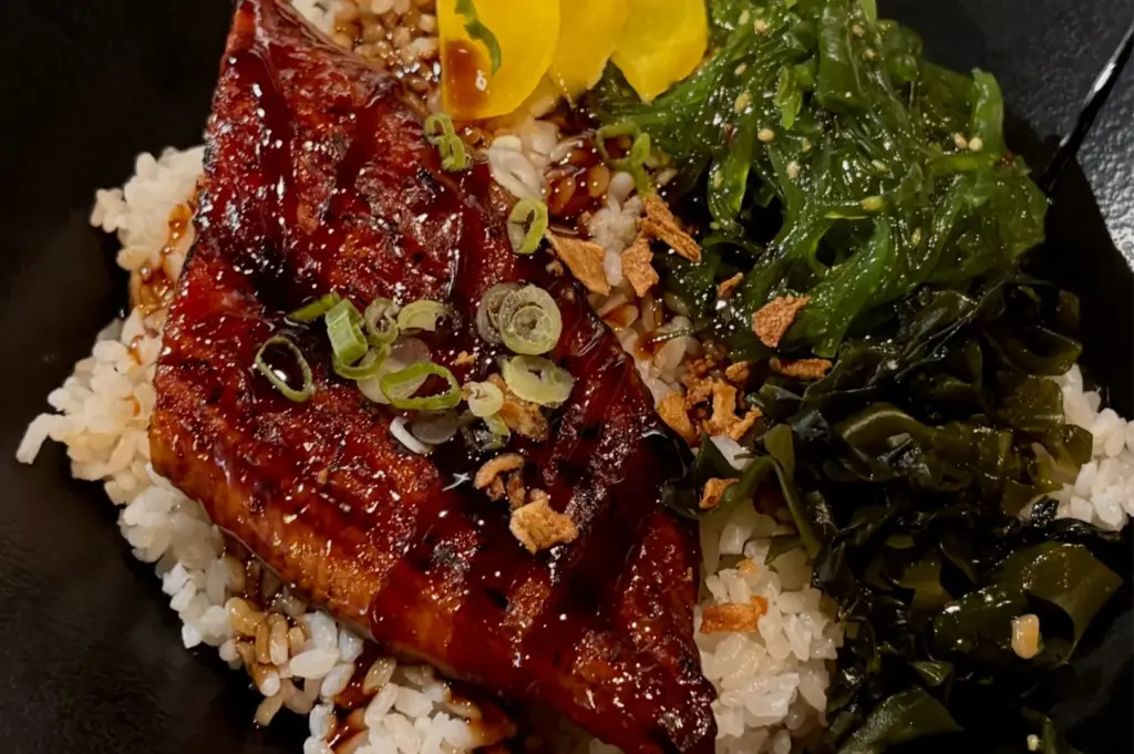A Japanese Unagi Don bowl featuring grilled eel with a sweet glaze, served over white rice with sides of seaweed salad and pickled ginger.
