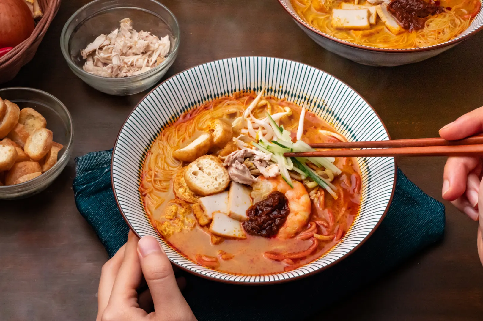 A close-up of two bowls of ramen, a spicy noodle soup, with ingredients like chili peppers, onions, and lemongrass arranged in a basket nearby.