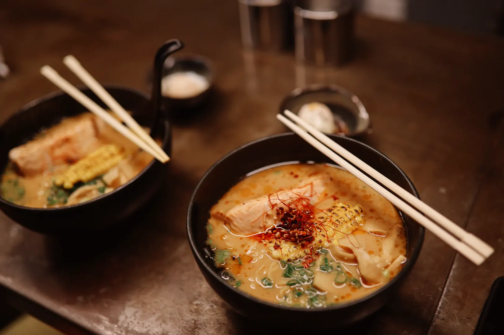 Two bowls of ramen noodles with chopsticks resting on top, garnished with meat, green onions, and chili flakes.