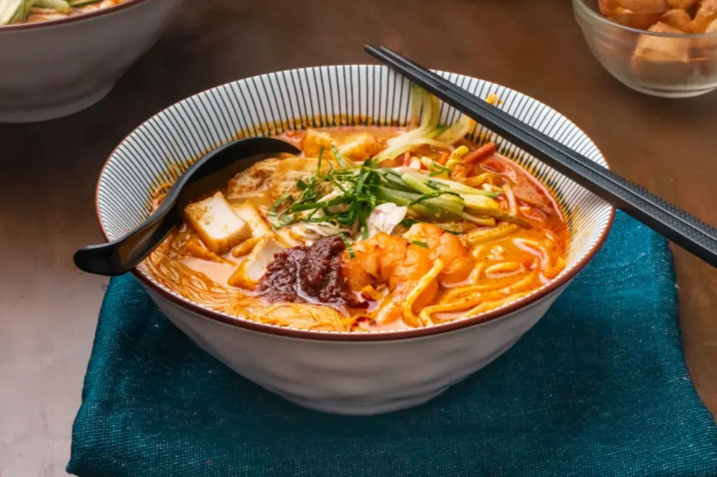 A bowl of ramen noodles, shrimp, tofu, and greens, served with chopsticks and a spoon on a teal cloth.