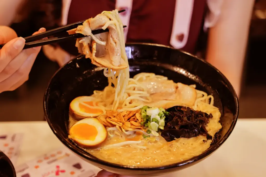 A black bowl of ramen with noodles, sliced pork, two halves of a soft-boiled egg, green onions, and black fungus, being lifted by chopsticks.