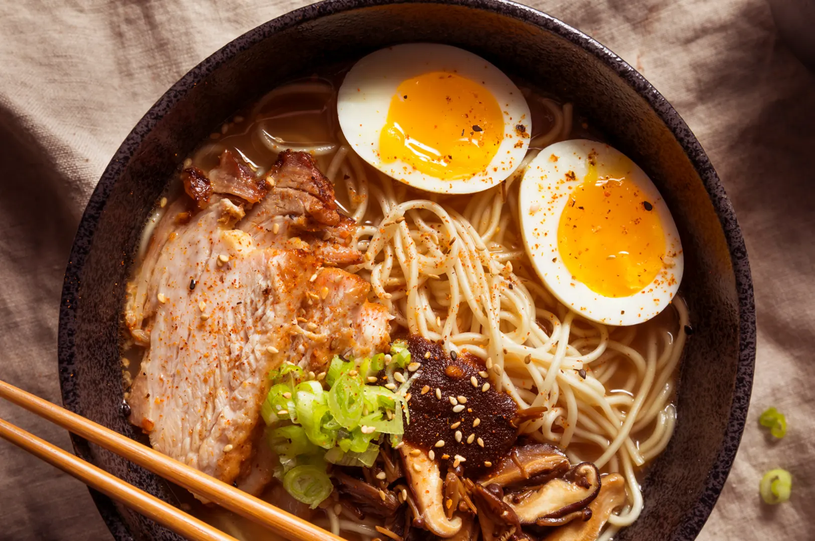 A bowl of ramen with noodles, sliced hard-boiled eggs, sliced pork, shiitake mushrooms, and chopped green onions, with chopsticks resting on the side.