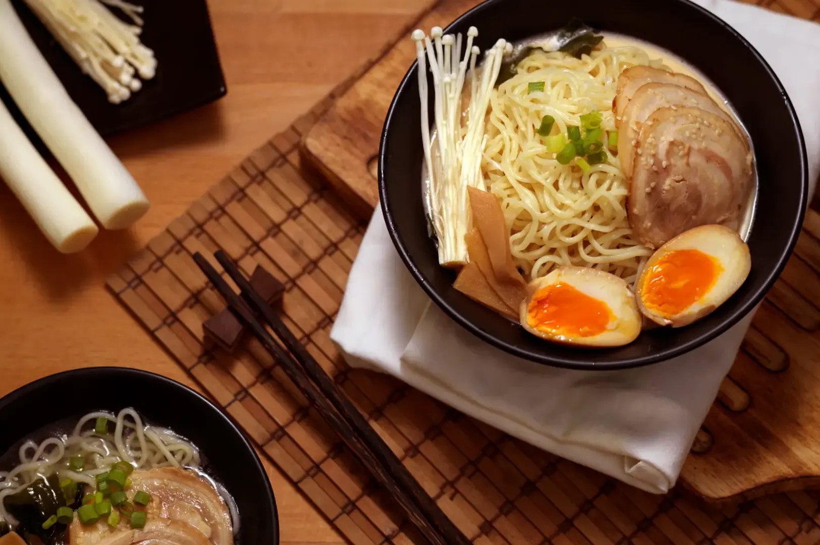 A bowl of ramen noodles with sliced pork, soft-boiled eggs, and enoki mushrooms, served with chopsticks and leeks on a wooden table.