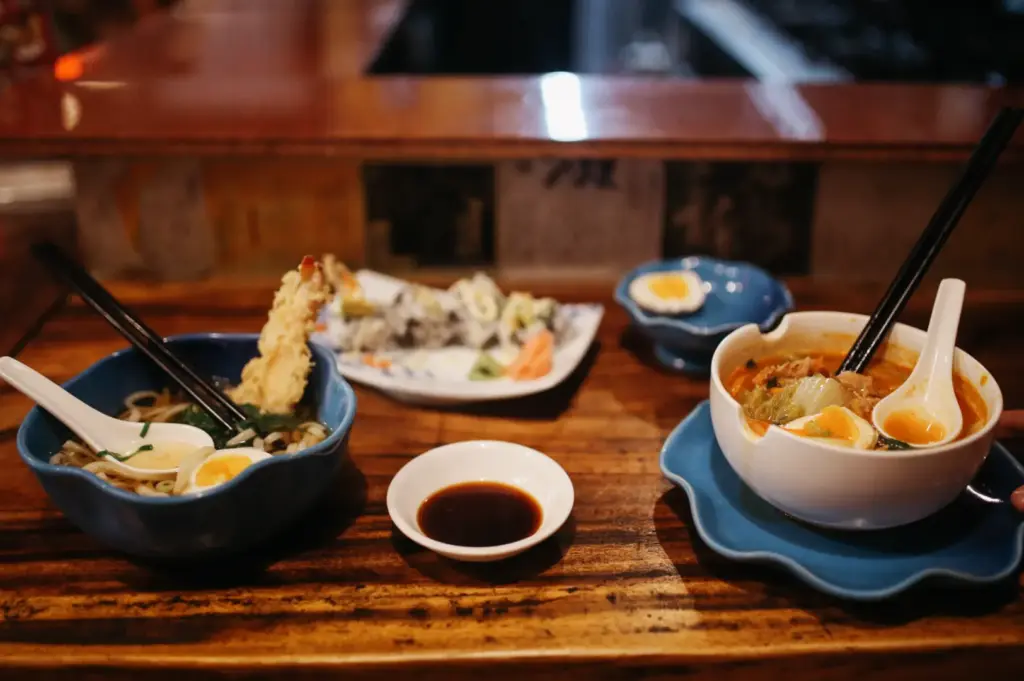 Two bowls of ramen and a plate of sushi rolls are served on a wooden counter.