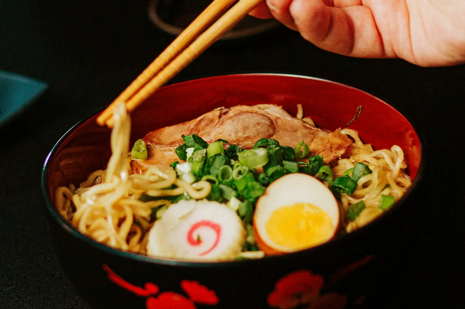 A hand uses chopsticks to lift noodles from a bowl of ramen topped with sliced pork, green onions, a hard-boiled egg, and a slice of narutomaki fish cake.