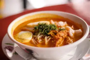 A bowl of laksa with noodles, shrimp, a hard-boiled egg, bean sprouts, and herbs in a rich orange broth.