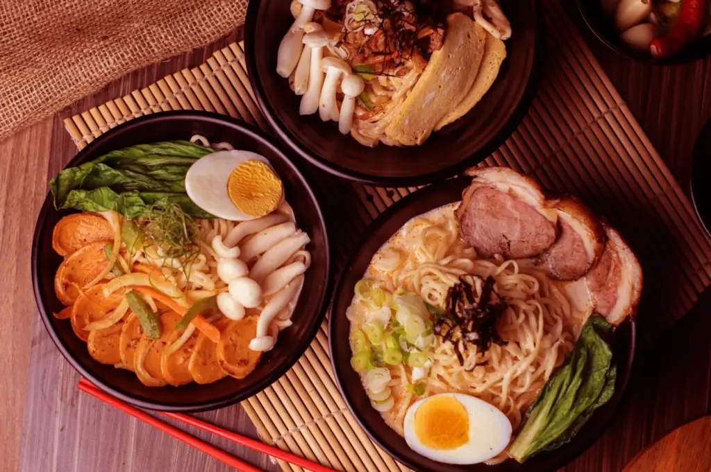 Three ramen bowls arranged on a bamboo mat, each with different toppings including pork, egg, and vegetables.