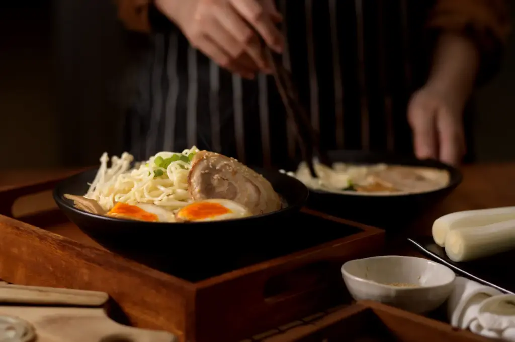 A bowl of ramen with noodles, sliced pork, a soft-boiled egg, and mushrooms, with a person in the background holding chopsticks over another bowl of ramen.