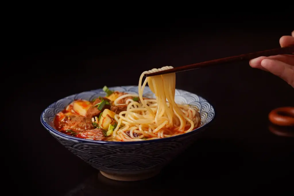 A hand uses chopsticks to lift noodles from a bowl of soup with meat and vegetables against a black background.