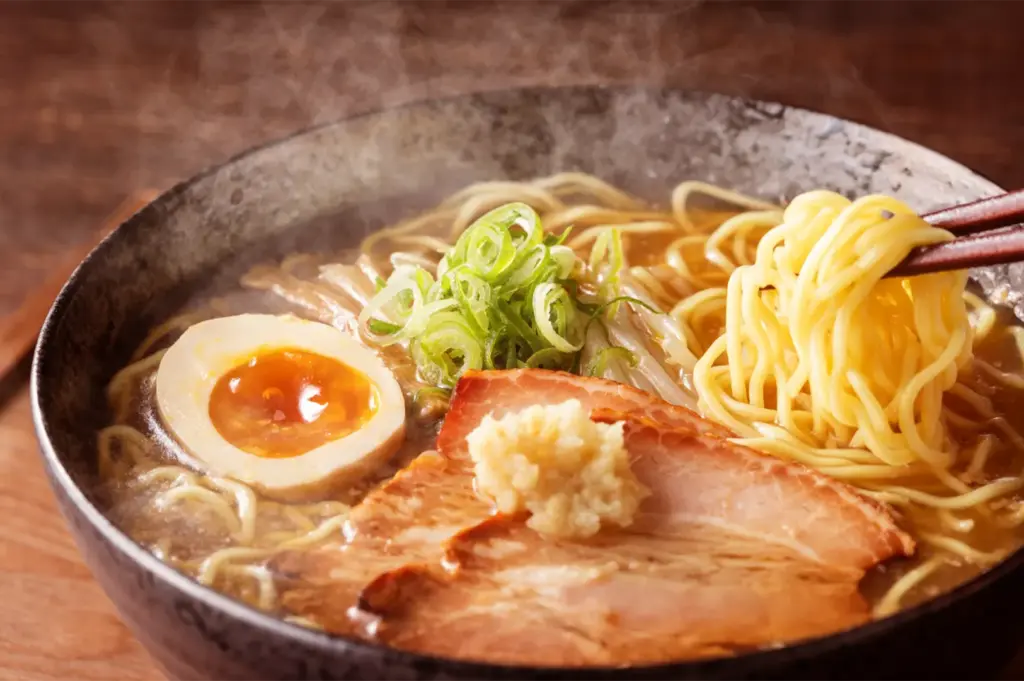 A steaming bowl of ramen noodles with sliced pork, a soft-boiled egg, and green onions, with chopsticks lifting a portion of noodles.