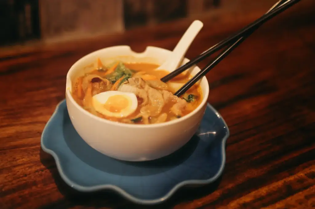 A bowl of ramen with sliced egg, meat, and vegetables, served with chopsticks and a spoon on a wooden counter.