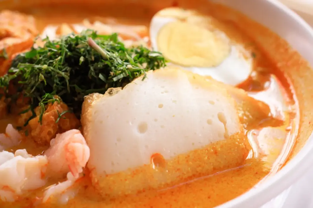 Close-up of Singaporean laksa noodle soup with rich orange coconut curry broth, topped with prawns, sliced fish cake, hard-boiled egg, and fresh herbs.