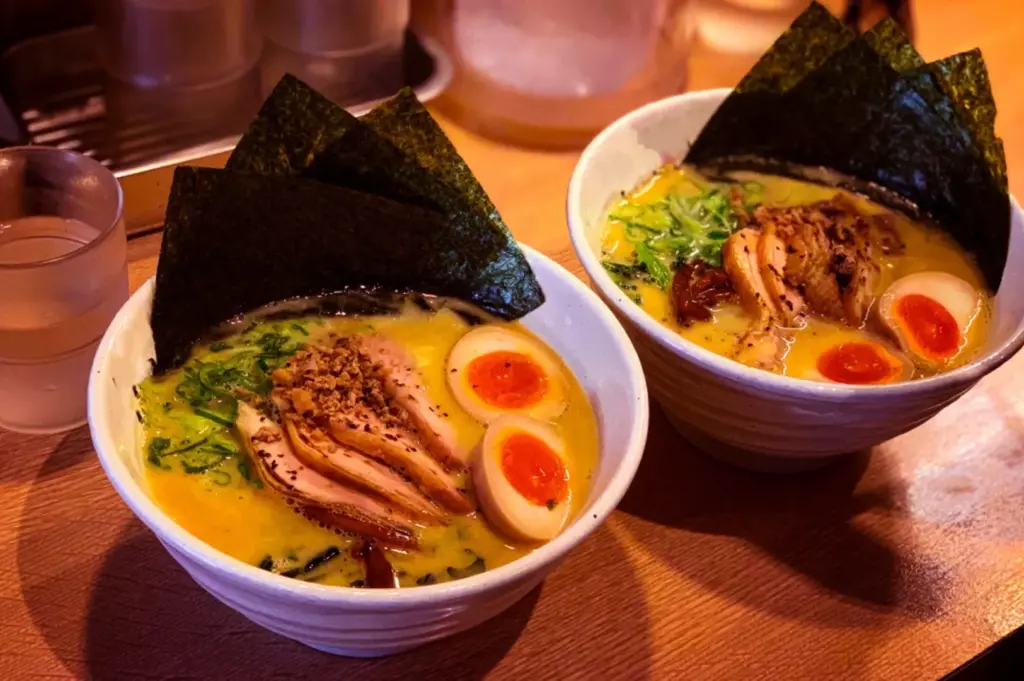 Two ramen bowls on a wooden table, topped with sliced pork, soft-boiled eggs, seaweed, and greens.