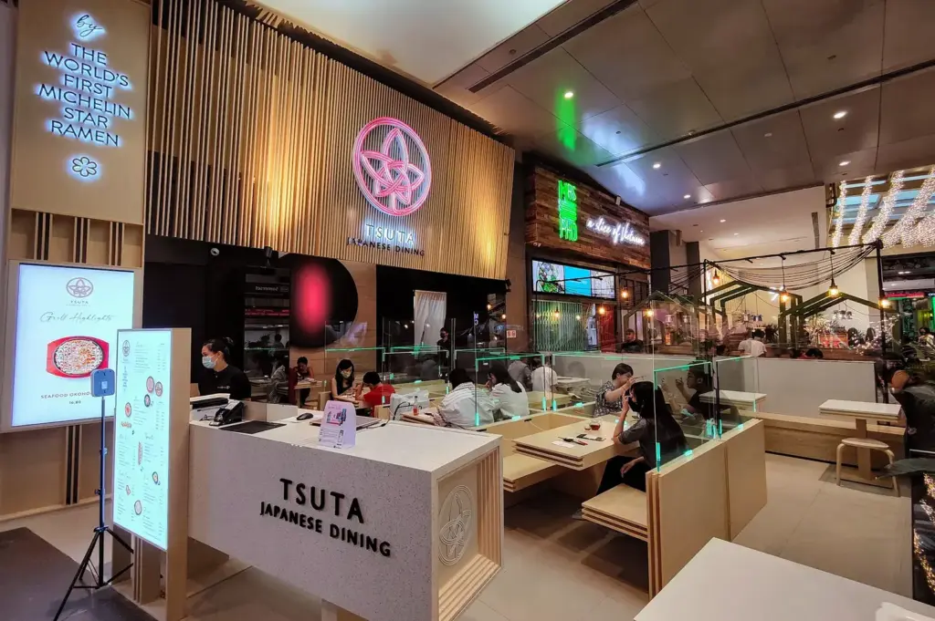 Interior view of Tsuta Japanese Dining restaurant, featuring modern wooden decor, a reception desk, and diners seated at tables.