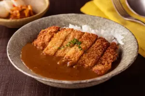 A bowl of Japanese curry with rice and sliced breaded pork cutlet, served with a side of pickles.