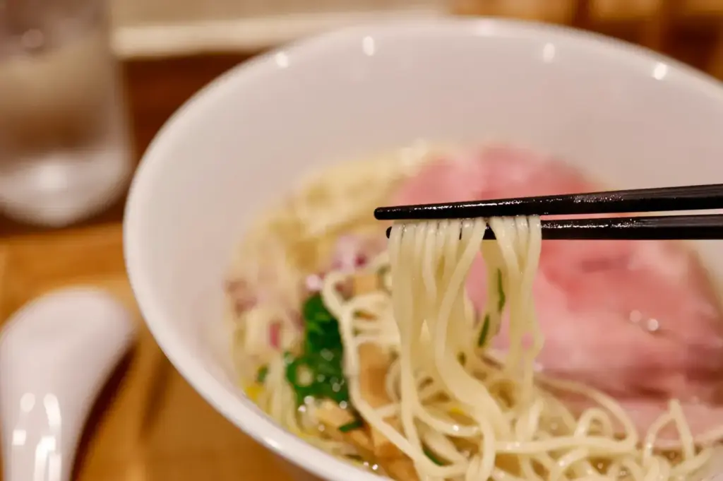 Chopsticks lifting noodles from a bowl of ramen with chashu pork.