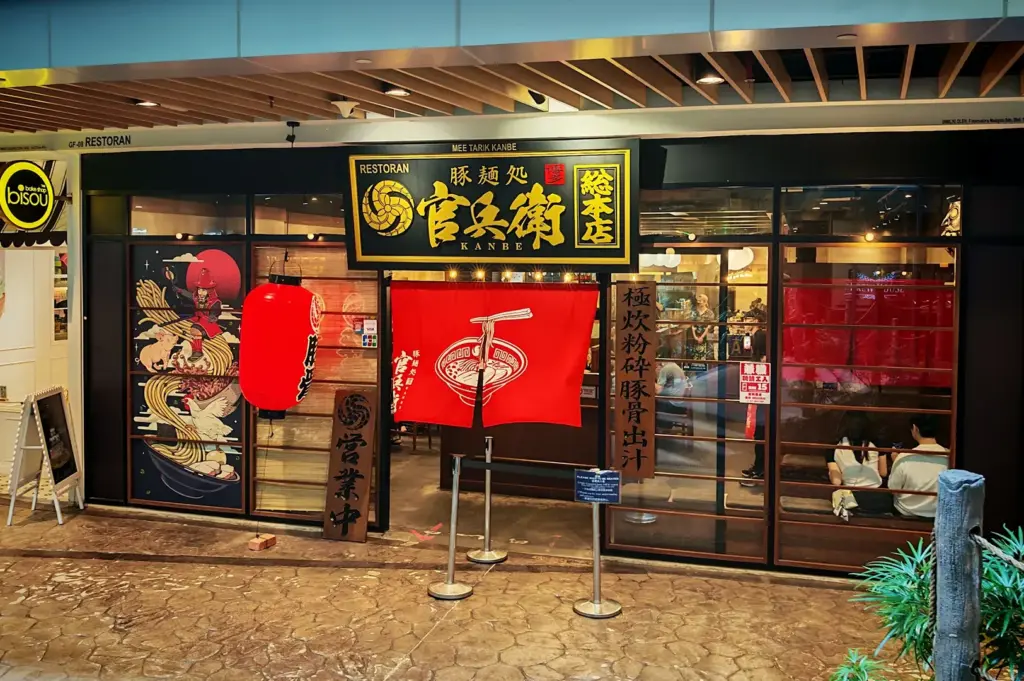 Kanbe Ramen restaurant entrance with red noren curtain, black signage with gold Japanese characters, dragon mural on left window, and queue stanchions outside.