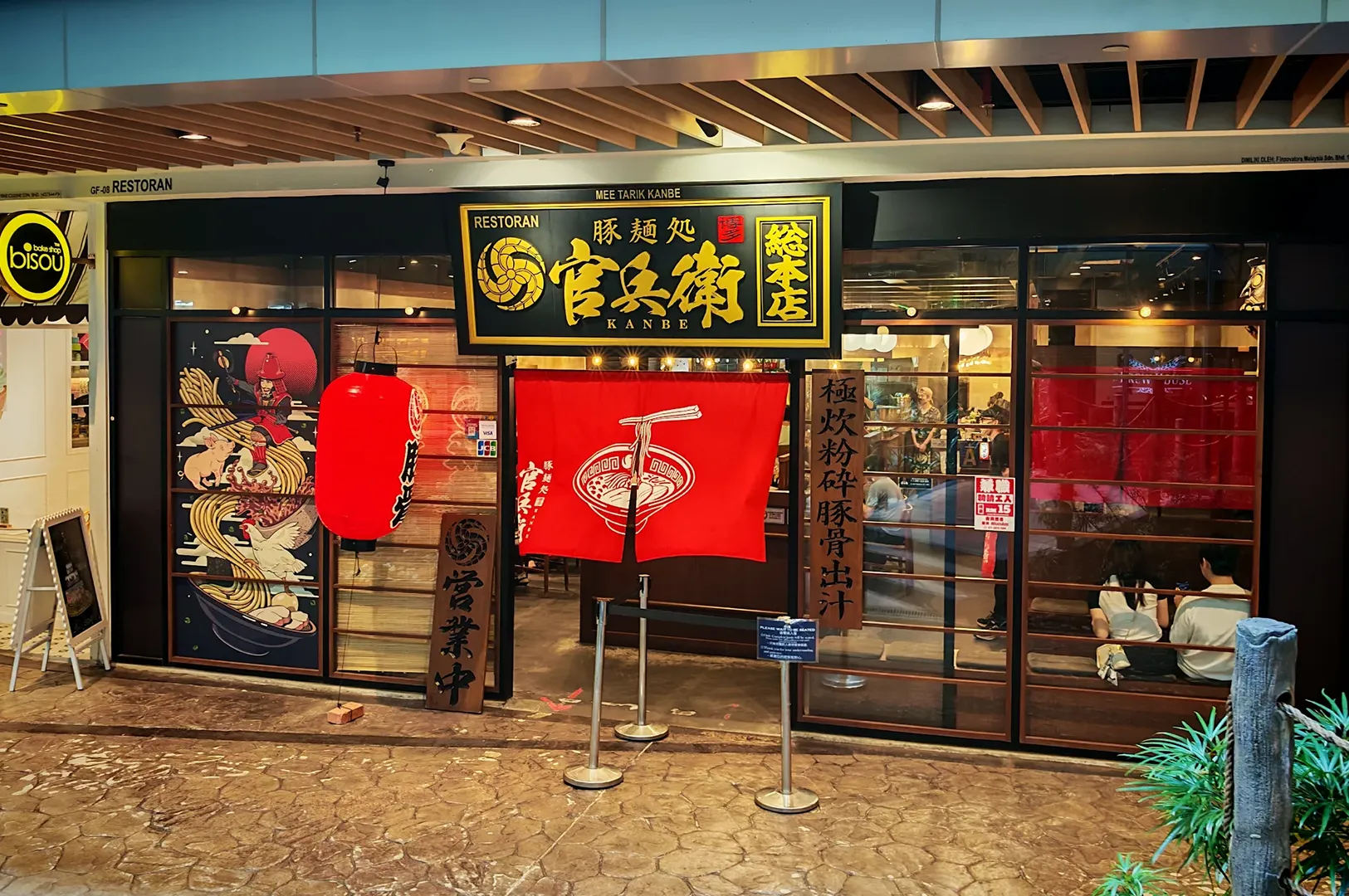 Kanbe Ramen restaurant entrance with red noren curtain, black signage with gold Japanese characters, dragon mural on left window, and queue stanchions outside.