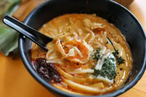 A bowl of laksa with noodles, shrimp, bean sprouts, and a dollop of chili paste, served with a spoon in each bowl. A green banana leaf package is visible in the background.