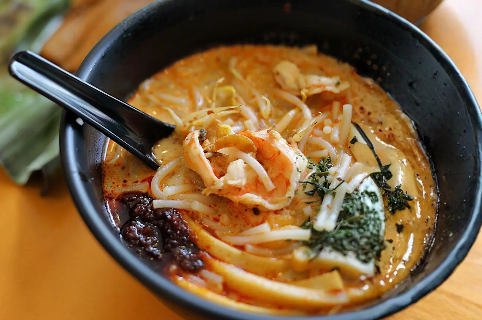 A bowl of laksa with noodles, shrimp, bean sprouts, and a dollop of chili paste, served with a spoon in each bowl. A green banana leaf package is visible in the background.