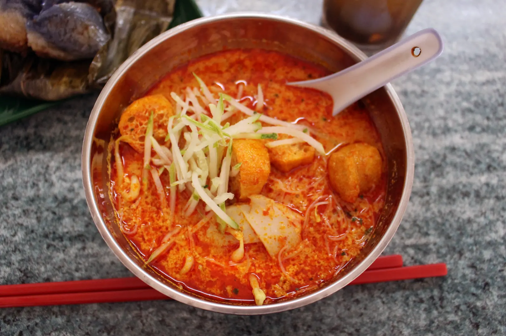 A close-up of a bowl of laksa, a spicy noodle soup, garnished with bean sprouts, fried tofu, and fish cakes. A spoon rests in the bowl, and red chopsticks lie beside it.