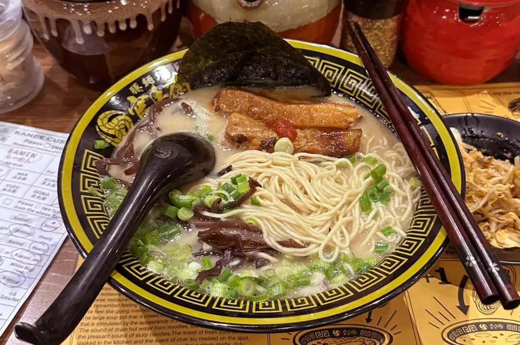 Ramen bowl with chashu pork, nori seaweed, wood ear mushrooms, and spring onions in creamy broth on yellow illustrated placemat with condiments in background.