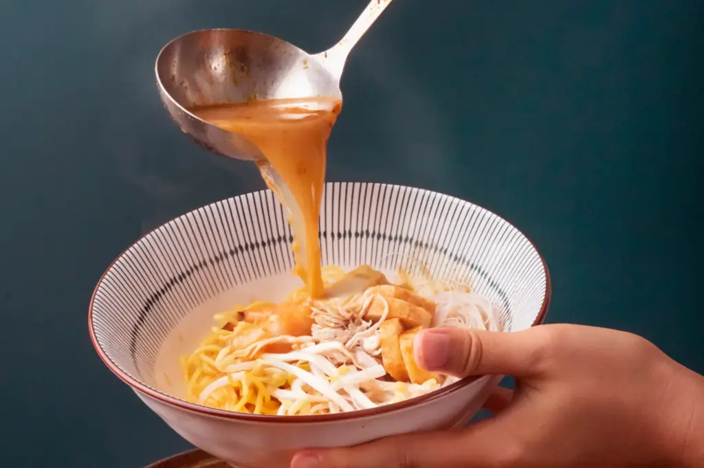A ladle pours a rich, golden broth over noodles and toppings in a patterned bowl, with a pot of the same broth visible below.