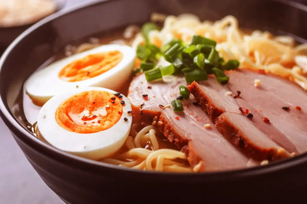 A bowl of steaming ramen with sliced pork, soft-boiled eggs, noodles, and chopped green onions.