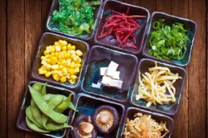 Ramen toppings in small bowls—corn, edamame, mushrooms, pickled ginger, scallions, bean sprouts, and seaweed.