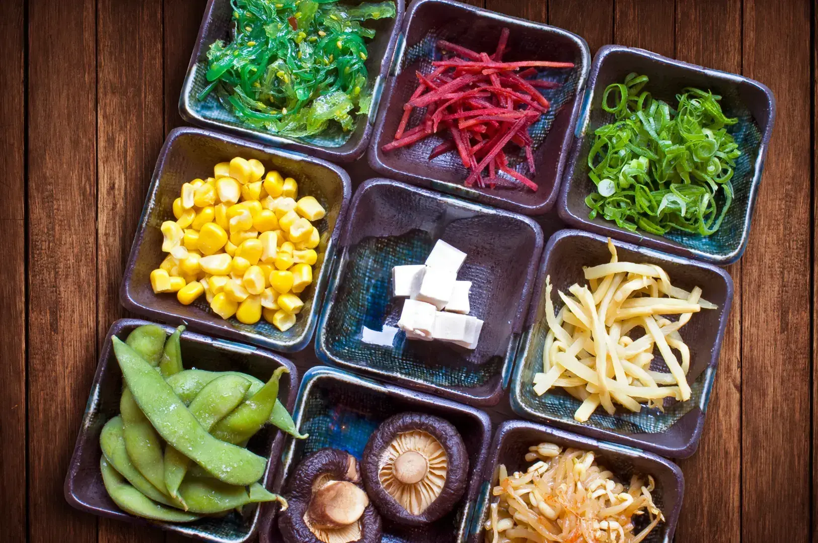 Ramen toppings in small bowls—corn, edamame, mushrooms, pickled ginger, scallions, bean sprouts, and seaweed.